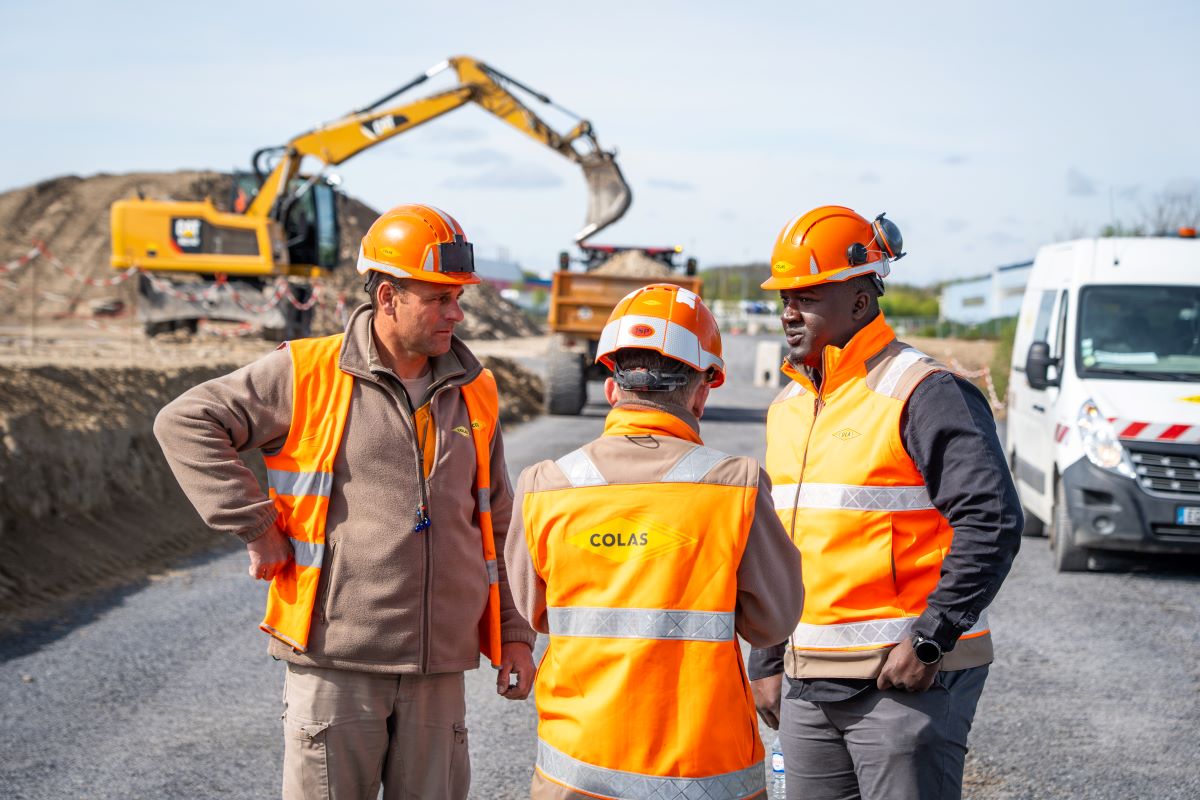 Trois agents Colas en discussion sur un chantier, engins de terrassement en arrière-plan.