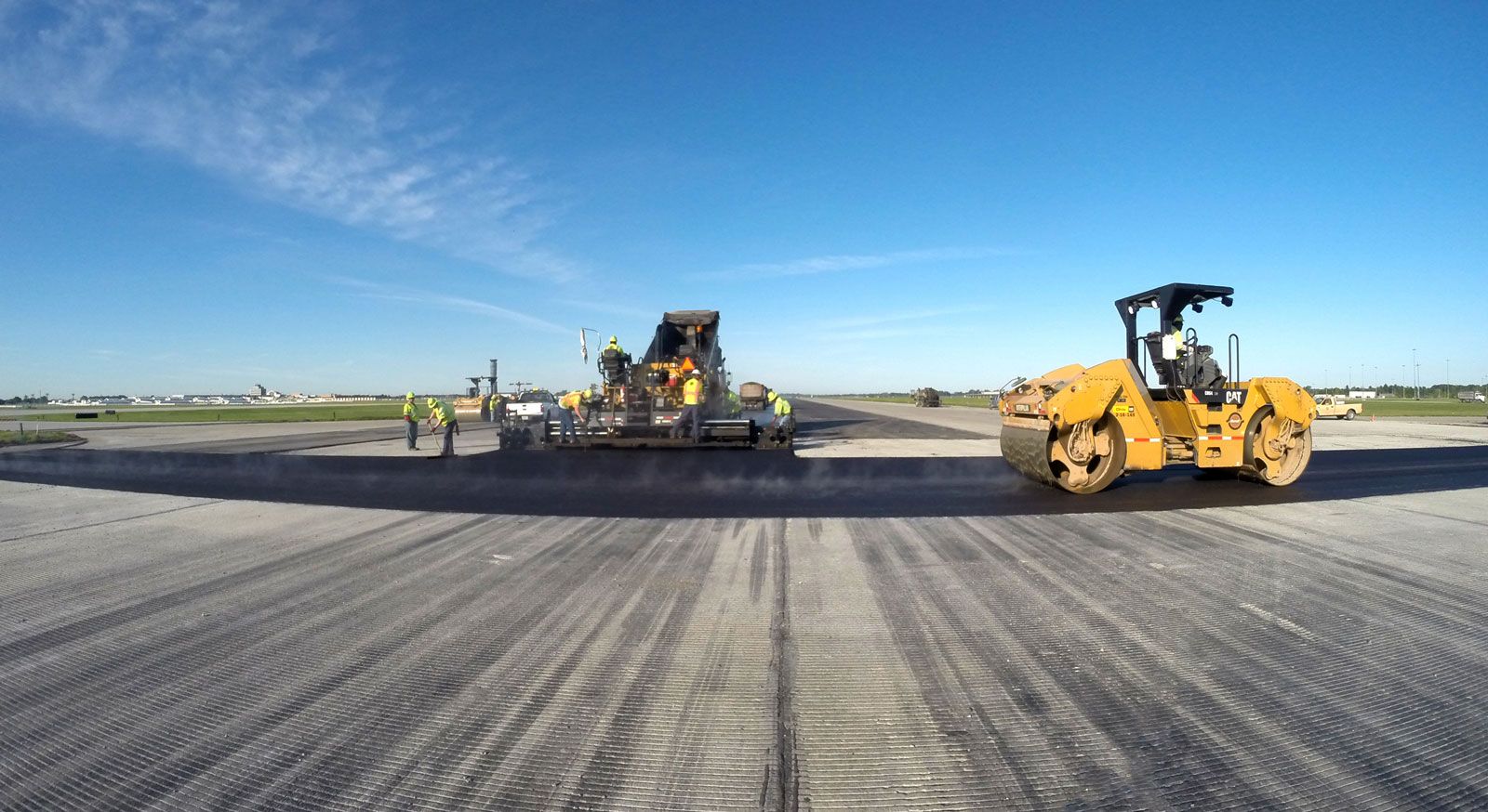 Travaux de construction sur une piste d'aéroport avec un rouleau compresseur et une équipe utilisant une machine, sous un ciel bleu.