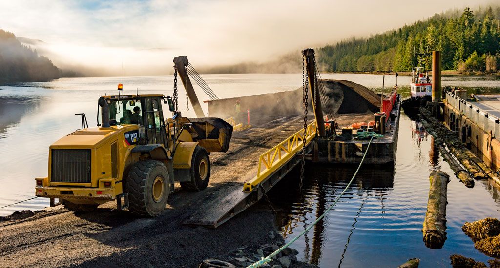Chantier de construction au bord de l'eau avec un bulldozer déplaçant de la terre sur une péniche, entouré de forêt et de montagnes embrumées.